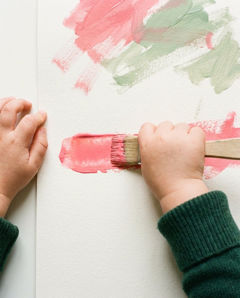Les mains d’un enfant étalent de la peinture rose au gros pinceau sur une grande feuille.